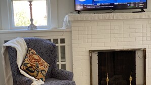 A living room with a blue armchair, a patterned throw pillow, a white fireplace, and a large flat-screen TV mounted above the fireplace.