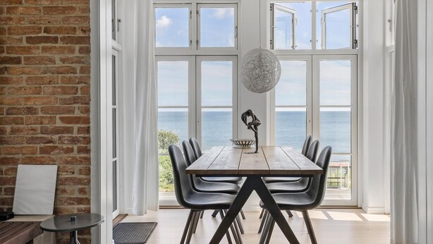 Un coin repas avec une table et des chaises en bois, de grandes fenêtres donnant sur l’océan, un luminaire suspendu et un mur en briques.