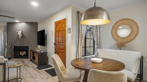 A dining area with a round wooden table, beige upholstered chairs, a large pendant light, and a wall-mounted mirror. - Sonoran Style 2BR/2BA w. Free Parking & Pool (Scottsdale)