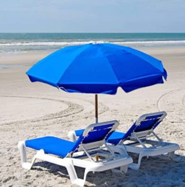 A beach with two white lounge chairs and a blue umbrella.