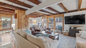 A spacious living room with a high ceiling, wooden beams, a large flat-screen TV, comfortable seating, and large windows.