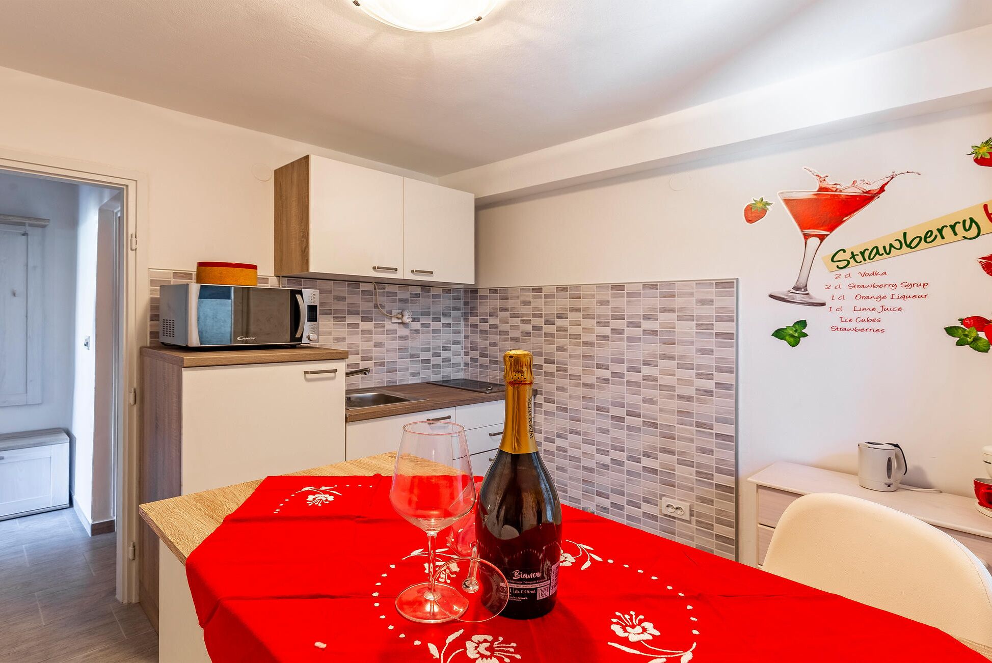 Une cuisine moderne avec une nappe rouge, une bouteille de vin et un verre. On peut voir une décoration murale représentant un cocktail fraise et une carte indiquant les ingrédients.