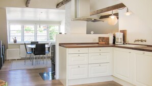 Una cucina moderna con mobili bianchi, un piano di lavoro in legno e illuminazione a sospensione. Si intravede un'area pranzo con tavolo e sedie attraverso una finestra.
