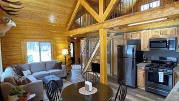 A spacious wooden cabin interior with a dining area, kitchen, and a staircase leading to an upper level.