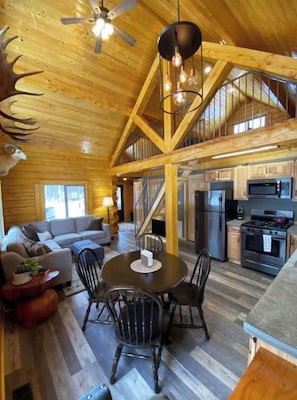 A spacious wooden cabin interior with a dining area, kitchen, and a staircase leading to an upper level. - Beautiful Comfy Alaskan Log Home 1 (Wasilla)