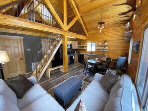 Cozy living area with a sofa, ottoman, and dining table, featuring a wooden staircase and large window.