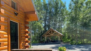 A log cabin with a wooden deck and a covered picnic area in a forest setting. - Beautiful Comfy Alaskan Log Home 1 (Wasilla)