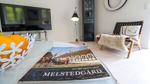 Ein Buch mit dem Titel 'Melstedgård' auf einem Tisch, ein Fernseher, eine Lampe, ein Stuhl und ein Ring an der Wand.