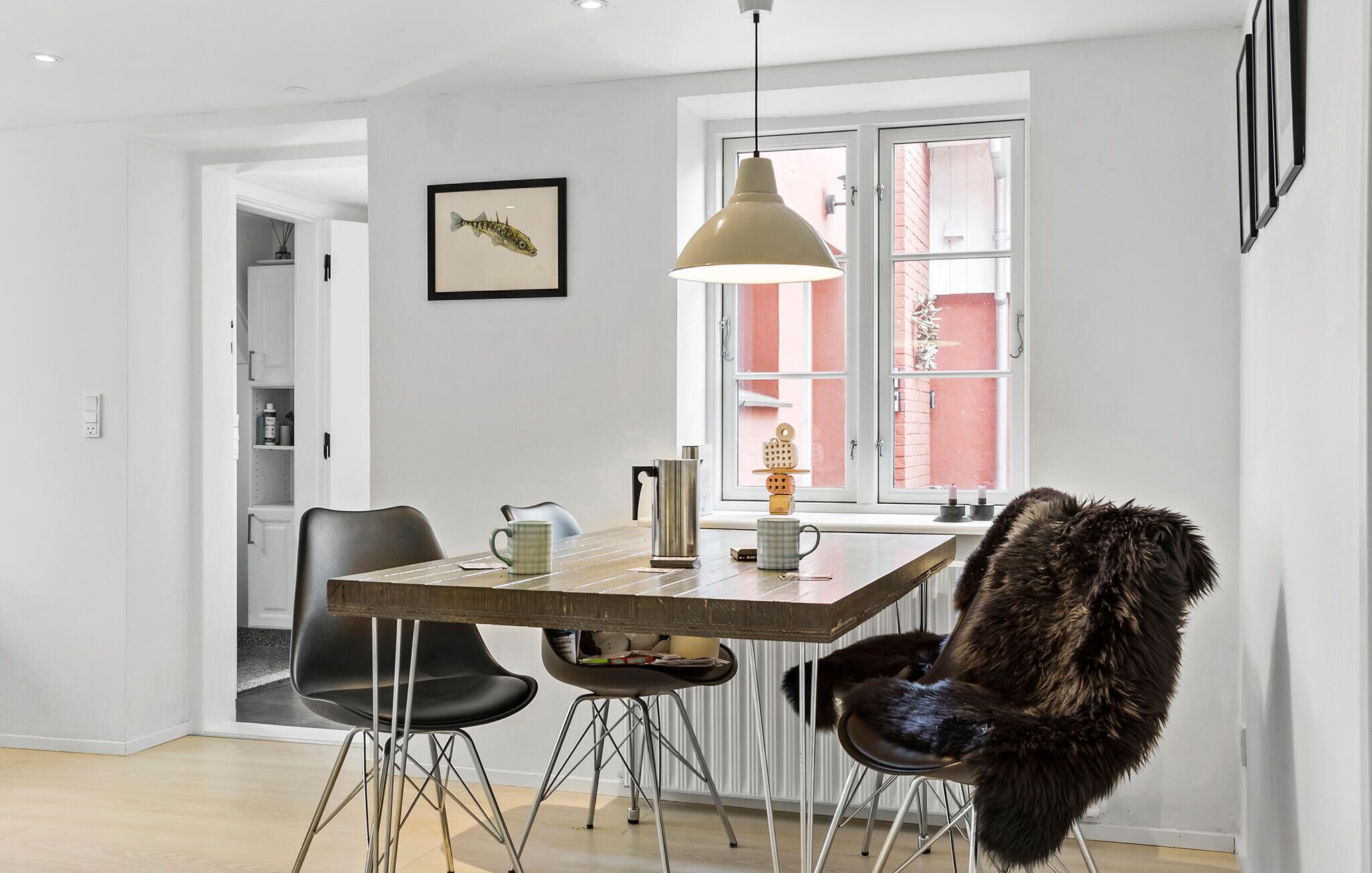 Un coin repas avec une table en bois, des chaises noires, un plaid en fourrure, une suspension et un tableau représentant un poisson, accroché au mur.