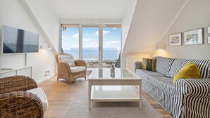 Ein modernes Wohnzimmer mit großem Fernseher, einem Korbstuhl, einem gestreiften Sofa und Blick aufs Meer.
