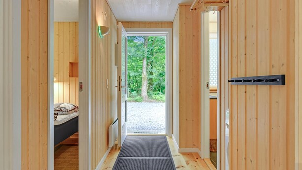 Un couloir intérieur en bois mène à une chambre à coucher équipée d’un lit et d’une table de chevet. On y trouve une porte avec une poignée, et la fenêtre offre une vue sur les arbres à l’extérieur.