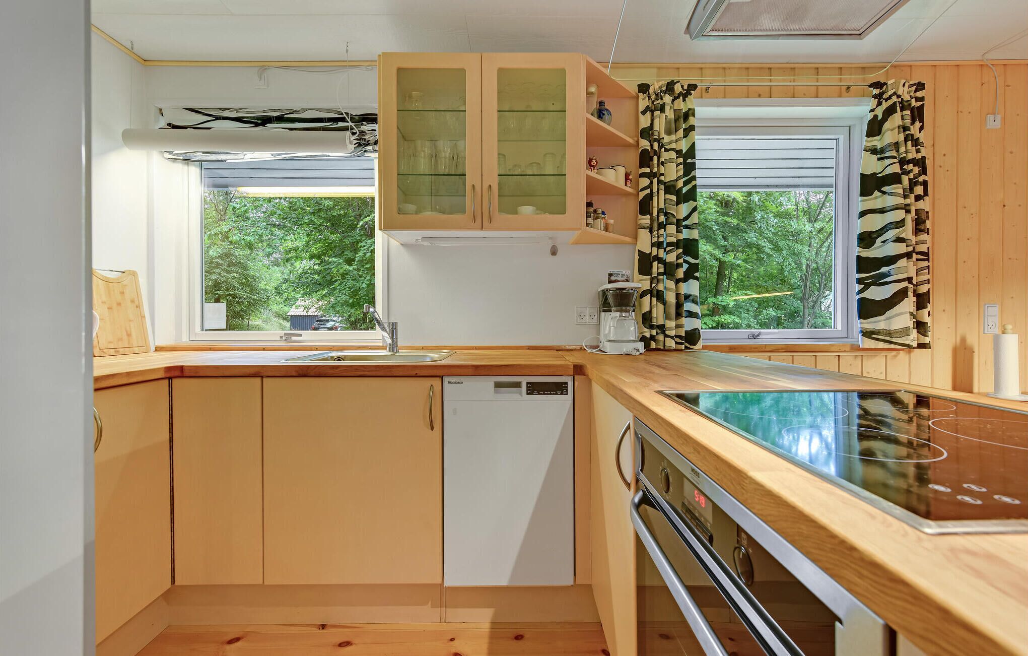 Une cuisine compacte avec des meubles en bois, un lave-vaisselle, une cuisinière et une fenêtre donnant sur un espace verdoyant.