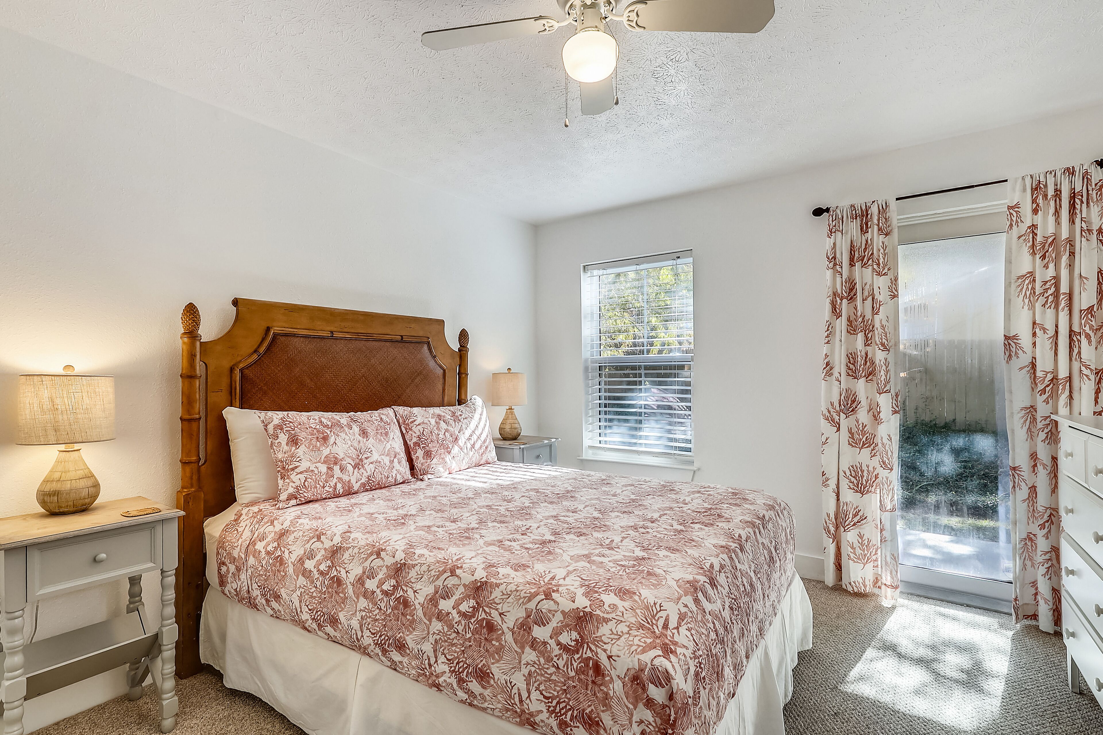 Quarto com cabeceira de madeira, cama com roupa de cama floral, mesa de cabeceira com abajur, janela com persianas e vista para o verde lá fora.