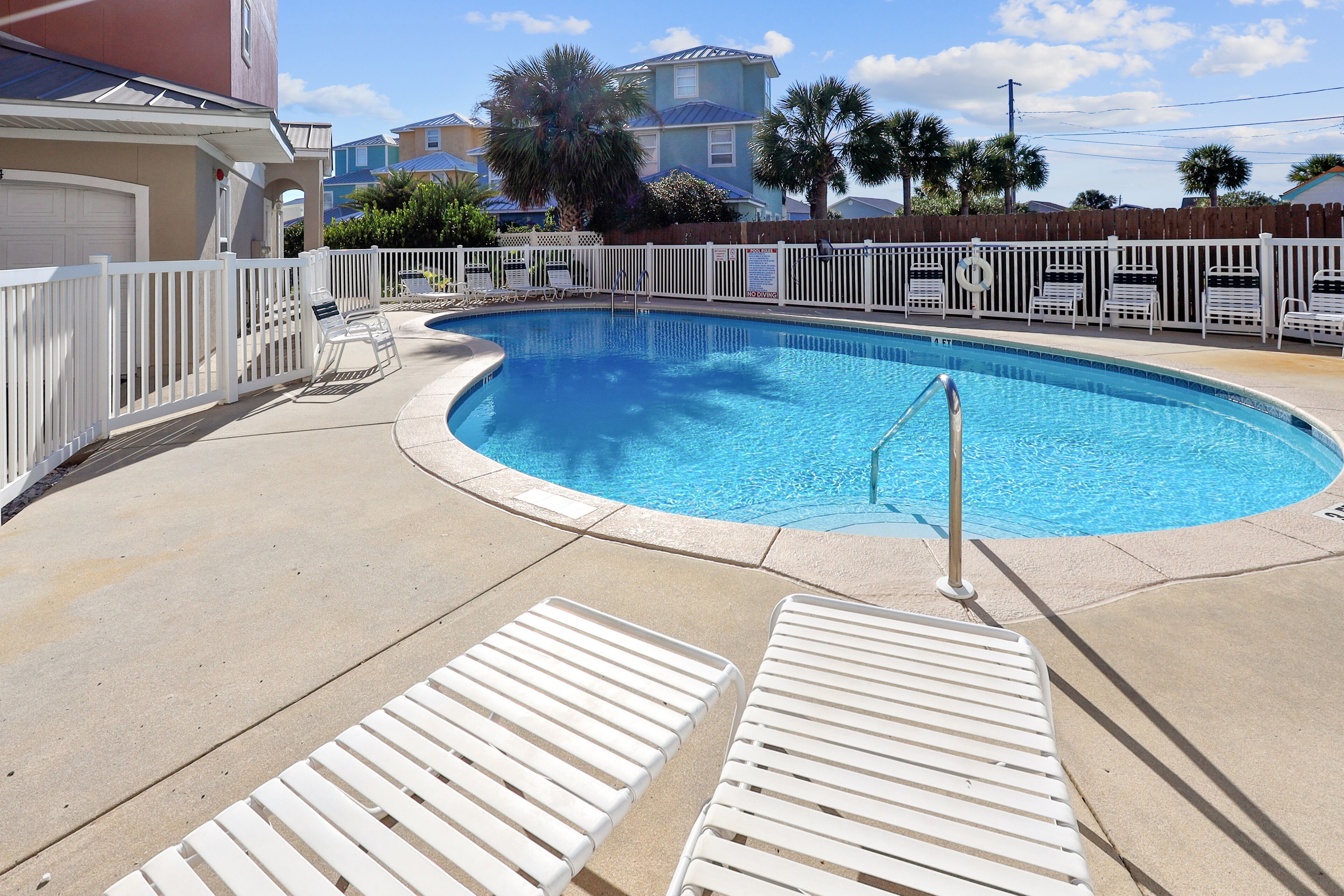 A swimming pool with a white fence, lounge chairs, and a clear blue sky.