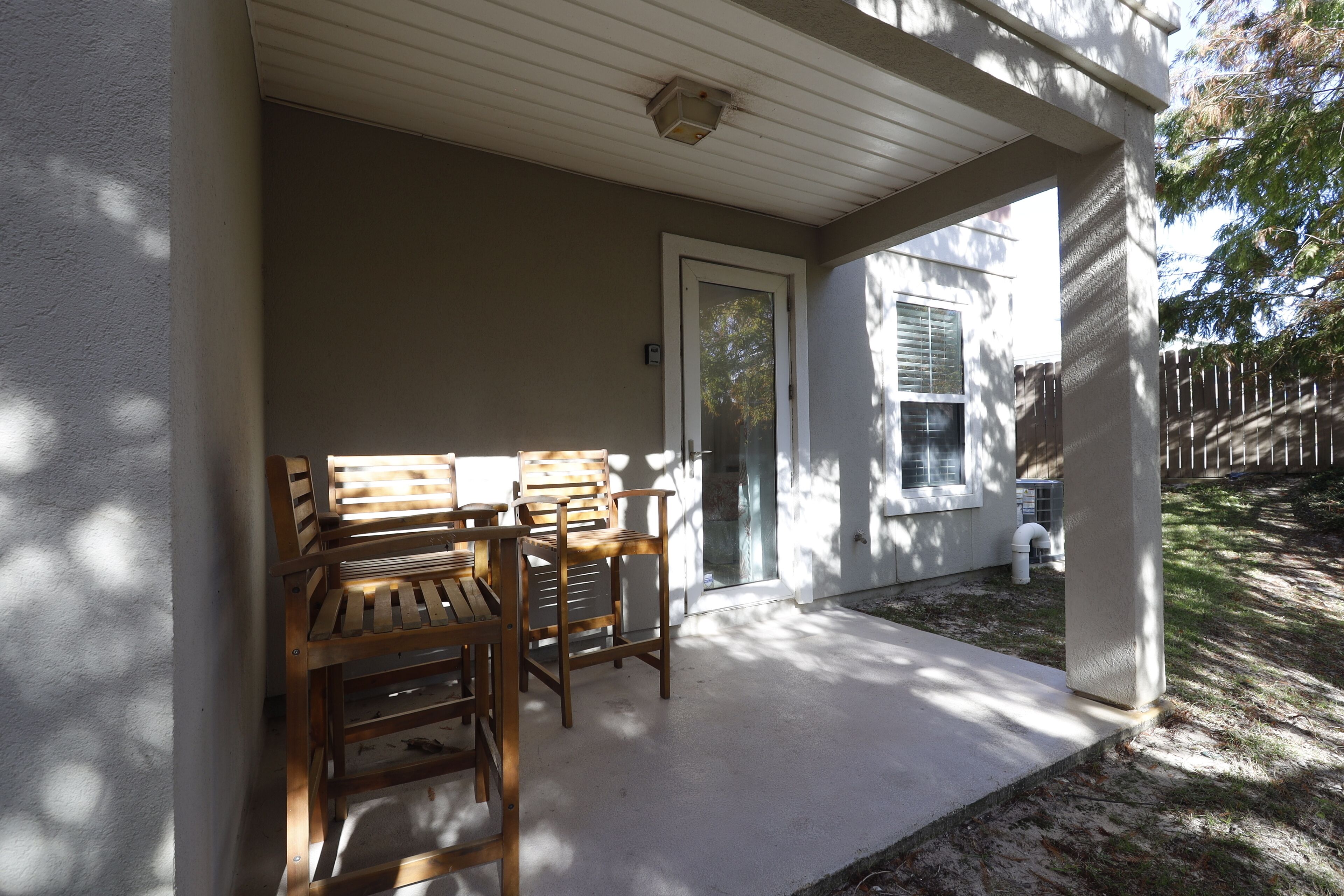 Une véranda abritée avec une table et des chaises, une porte et une vue sur un jardin avec une clôture et des arbres.