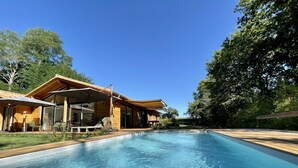 Un espace moderne aménagé autour d’une piscine, avec une maison en bois, des sièges d’extérieur et une piscine à l’eau d’un bleu limpide.
