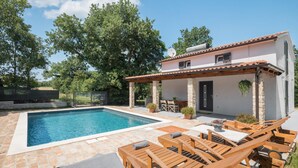 A swimming pool with lounge chairs and a patio area, surrounded by trees and a building with a tiled roof. - Pet friendly home in Krnicari (Krnicari)
