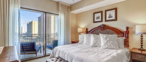 A hotel room with a large bed, a wooden headboard, a bench, a nightstand, and a view of the city through a sliding door.