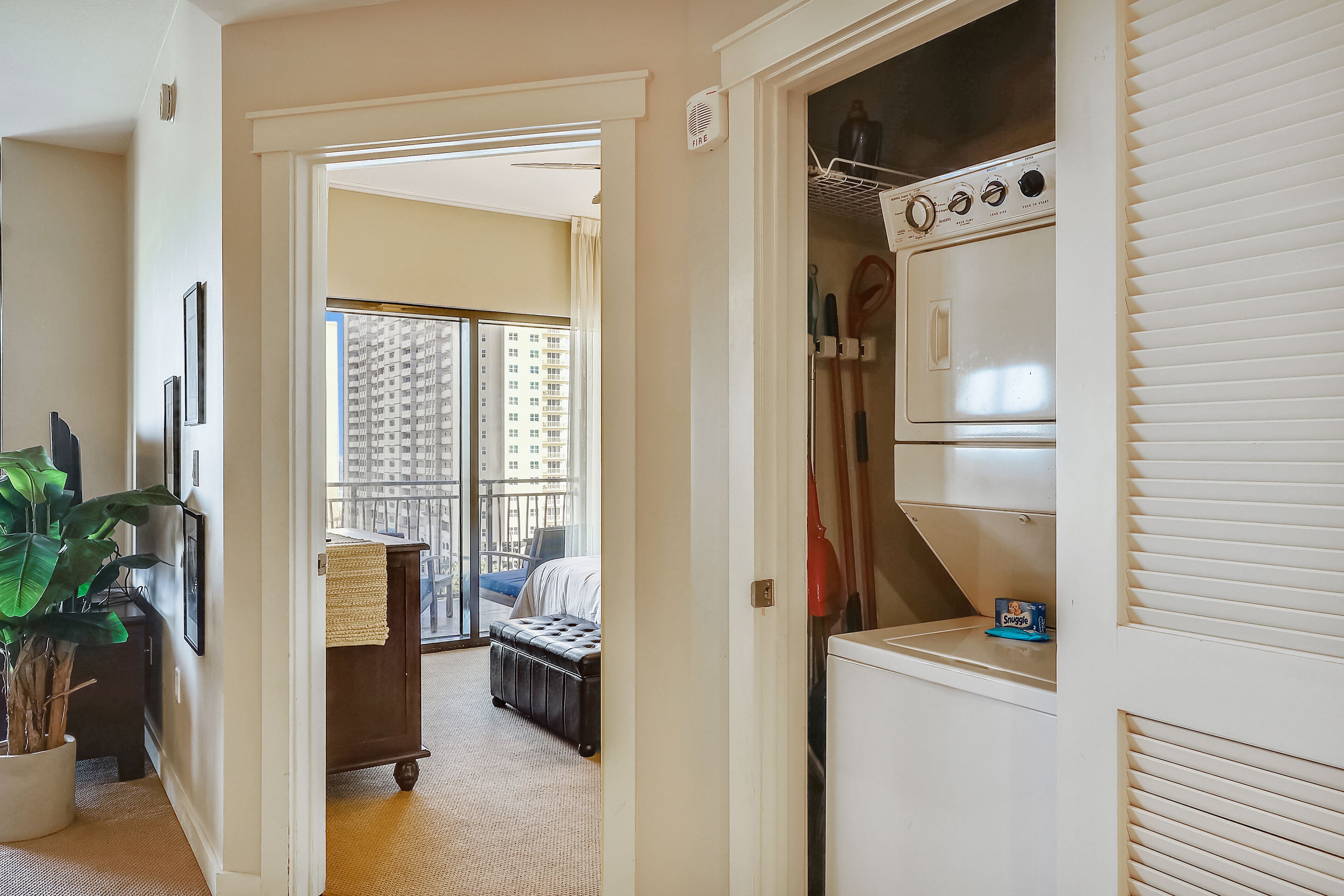 A hotel room with a large window, a bed, a closet with a washing machine, and a potted plant.