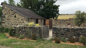 Une ferme en pierre avec une porte en bois, entourée d’un mur de pierre et d’une zone herbeuse où poussent de petites plantes.