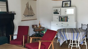 Un salon avec un plancher en bois, un fauteuil rouge, une table à manger recouverte d’une nappe à rayures et une lampe suspendue.