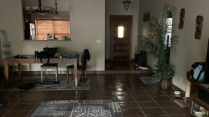 Een ruime woonkamer met een betegelde vloer, een eettafel met stoelen, een keuken met een raam, en een gang naar een andere kamer.