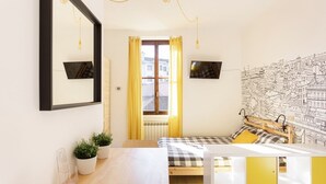 Ein gemütliches Zimmer mit Bett, Tisch, Stühlen, Spiegel, Fernseher und einem Fenster mit Vorhängen.
