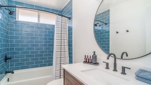 Un baño con azulejos azules, ducha, inodoro, lavamanos con espejo y artículos de aseo.