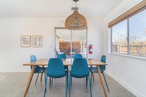 A modern dining area with a wooden table and teal chairs, a woven pendant light, and a large window with a view of the outdoors. - JT Agave Oasis | 3BR + Game Room + Cowboy Pool (Joshua Tree)