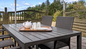 Ein Tisch im Freien mit Teekanne, Tassen und Tellern auf einem Tablett, umgeben von Korbstühlen und einem Holenzaun.