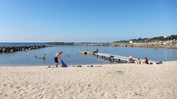 Ein Strand mit einem Steg, der sich in ruhiges Wasser erstreckt, Menschen liegen in der Sonne und eine felsige Küste mit Gebäuden im Hintergrund.