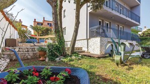 Ein Wohngebäude mit einer Stein-Fassade, einem Balkon mit Metallgeländer und einem Gartenbereich mit Kübelpflanzen und Blumen.