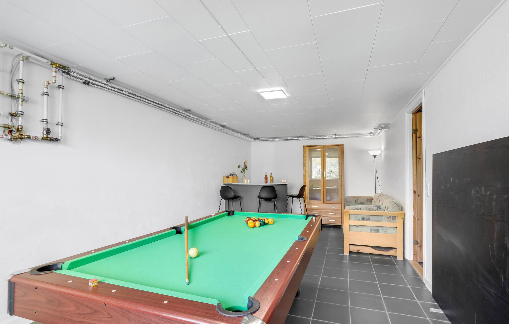 A pool table in a room with a kitchenette area, a wooden cabinet, and a door leading to another room.