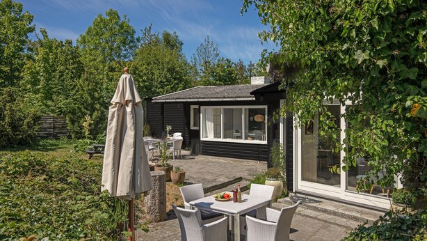 A small, dark wooden cottage with a patio area featuring a table and chairs, surrounded by lush greenery. - 3 bedroom amazing home in Nexø (Nexø)