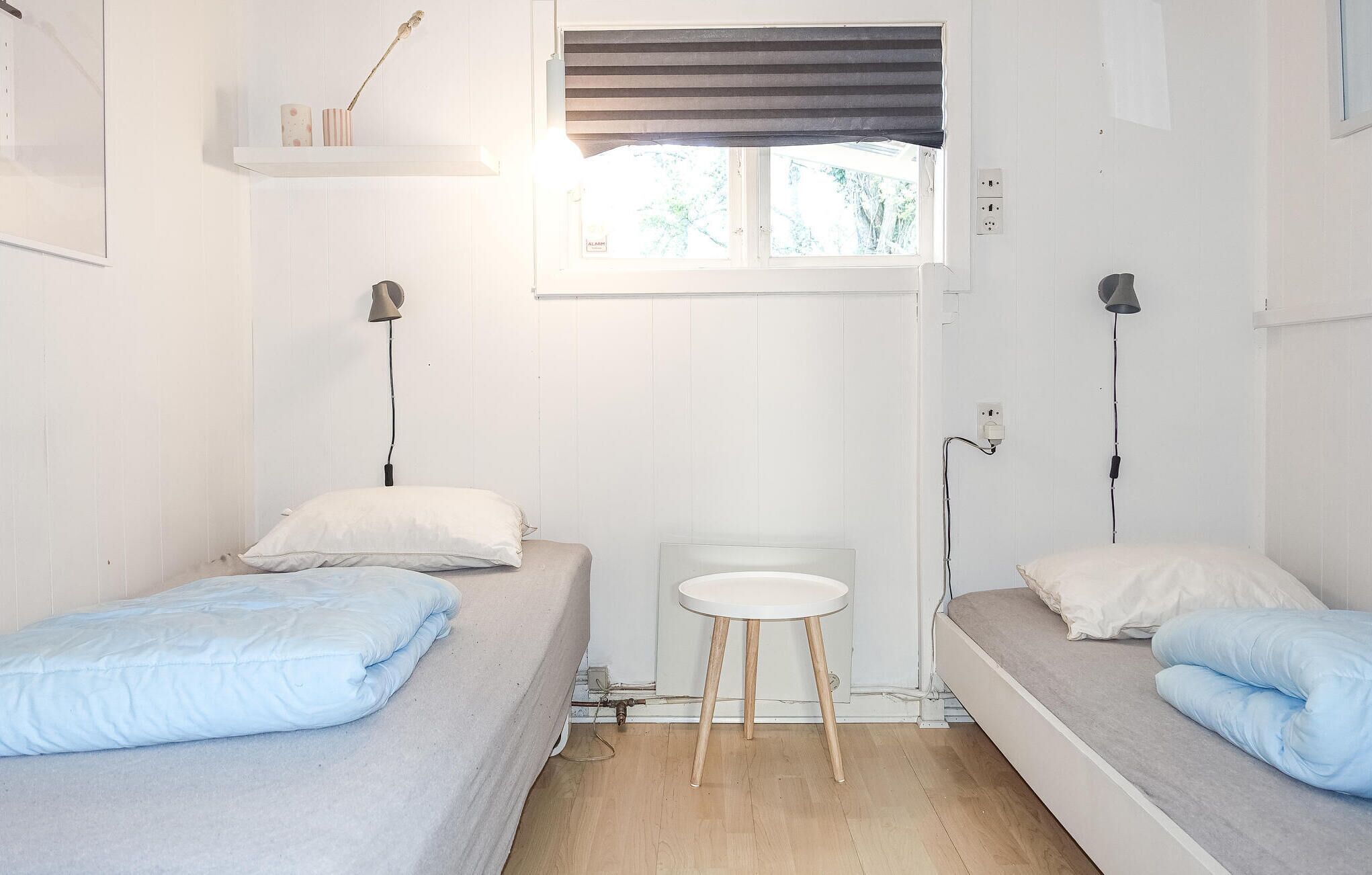 A small, well-lit room with two single beds, a small table, and a window with a striped blind.