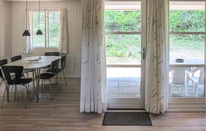 A dining area with a table and chairs, large windows with curtains, and a view of greenery outside.