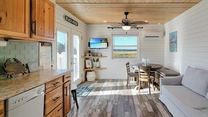 A compact kitchen with wooden cabinets, a dining area with a table and chairs, a sofa, and a large window offering a view of the outdoors. - BAY-CAY Getaway, Beaching, Casinos, Swim, Grill, Chill, Memories! (Bay St. Louis)