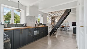 Une cuisine moderne avec des meubles de couleur sombre, un plan de travail en bois et un grand îlot central. Un escalier orné d’un motif en filet de corde mène à un espace repas comprenant une table et des chaises.