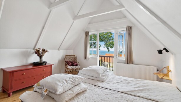 Une chambre avec un plafond en pente, un lit recouvert de linge de lit blanc, une commode rouge, un fauteuil avec des coussins et une vue sur la mer par la fenêtre.