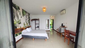 A bedroom with a bed, a desk, and a chair, a small table with a plant, and a wall with a tropical mural. - Queen Suite (Montezuma)
