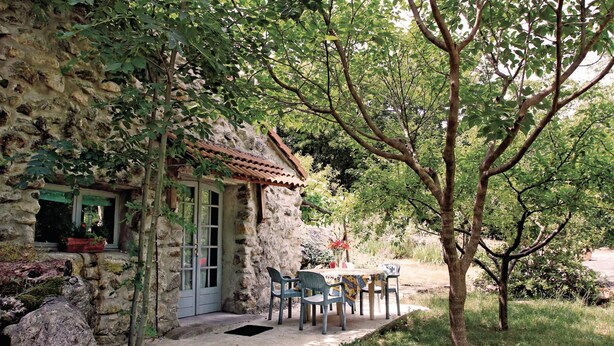 A stone building with a tiled roof, a door, and a window. There is a table with chairs outside, and a tree in front.