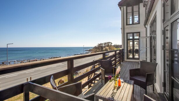 En balkon med udsigt over stranden og havet, en træterrasse og en bygning med store vinduer.