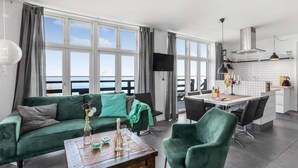En moderne stue med en grøn sofa, et træbord og et spiseområde med bord og stole. Et fjernsyn er monteret på væggen, og der er en balkon med udsigt.