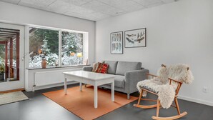 Ein modernes Wohnzimmer mit einem weißen Tisch, einer grauen Couch und einem Schaukelstuhl. An der Wand befinden sich gerahmte Bilder und ein Fenster mit Blick auf schneebedeckte Bäume.