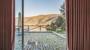 A view from a window with curtains, featuring a cobblestone patio, a metal railing, and a mountainous landscape in the background.