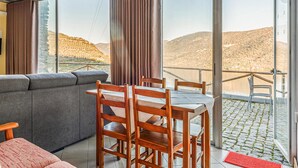 A modern room with a grey sofa, wooden chairs, a dining table, and large windows offering a view of a mountainous landscape.