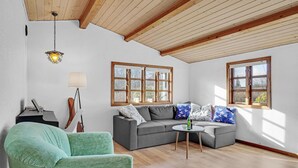 Un soggiorno con un divano grigio, una poltrona verde e un tavolino rotondo. Sono presenti due finestre con stipiti in legno, un lampadario a sospensione e una lampada da terra.