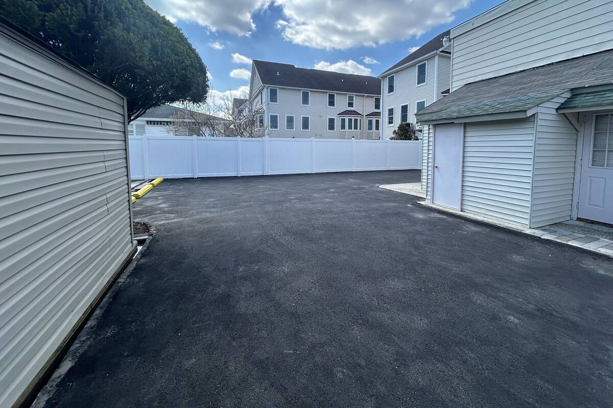 A parking lot with a white fence, a building with a garage, and a house in the background.