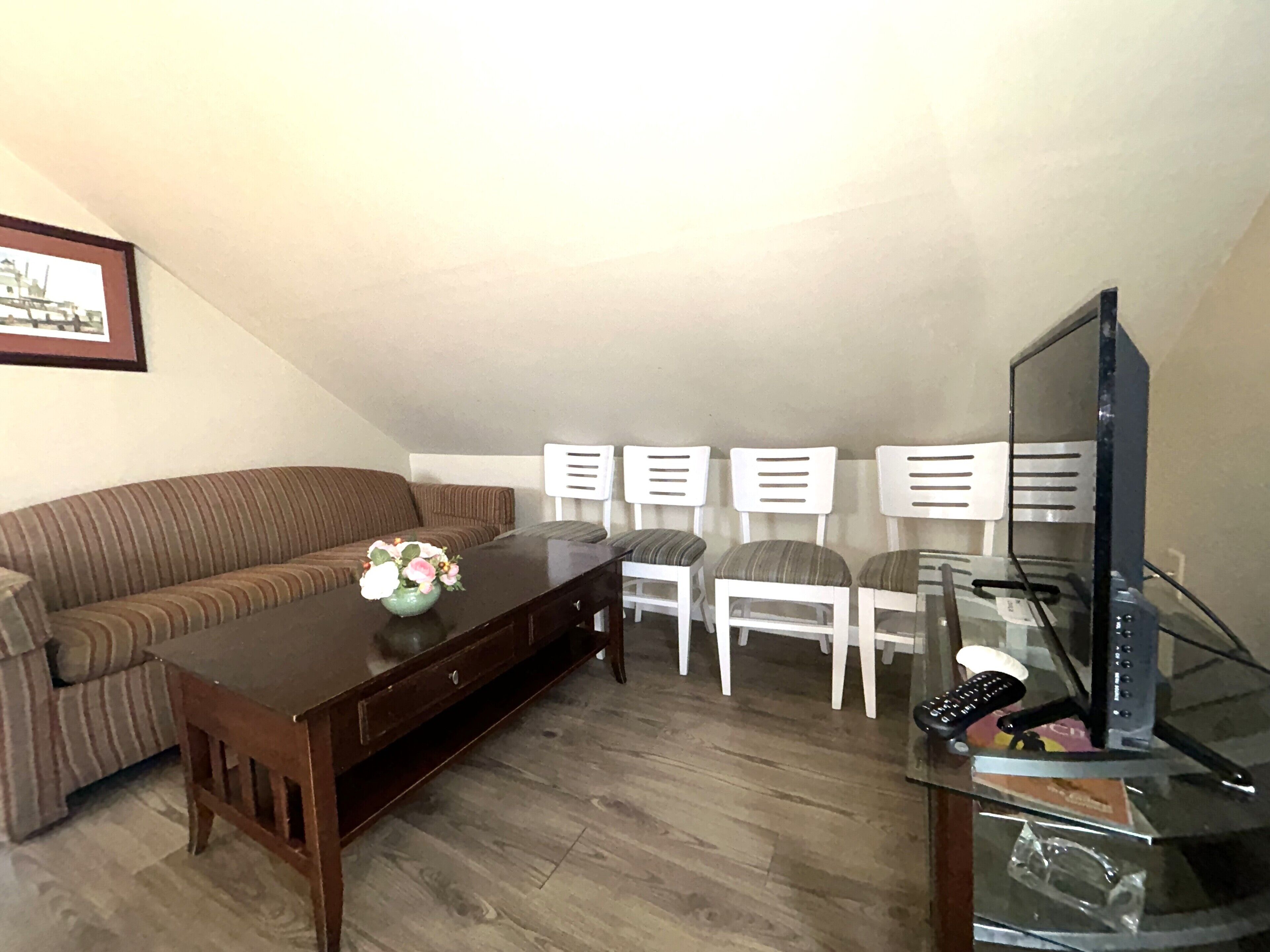 Ein Wohnzimmer mit einem braunen Sofa, einem Holztisch und einer TV-Konsole aus Glas mit einem Flachbildfernseher. Es gibt drei weiße Sessel und ein gerahmtes Bild an der Wand.