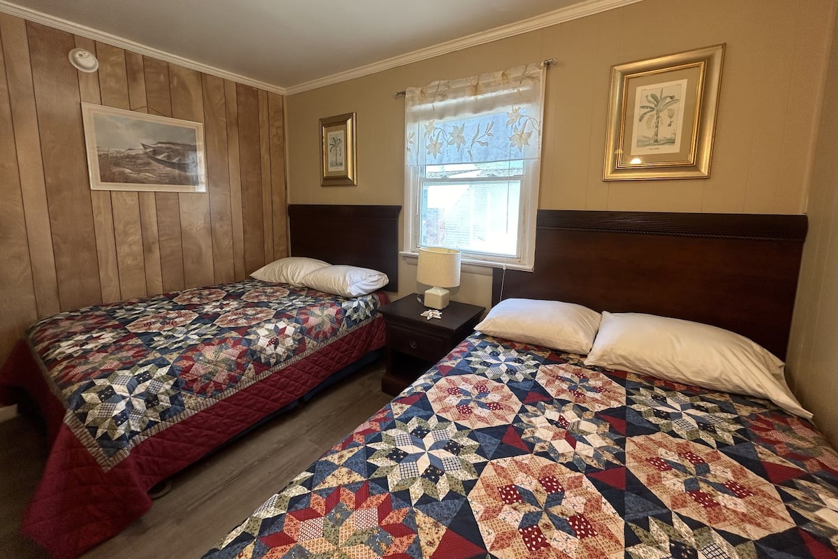 A room with two beds, each with a quilt, a nightstand with a lamp, and a window with a view of trees.